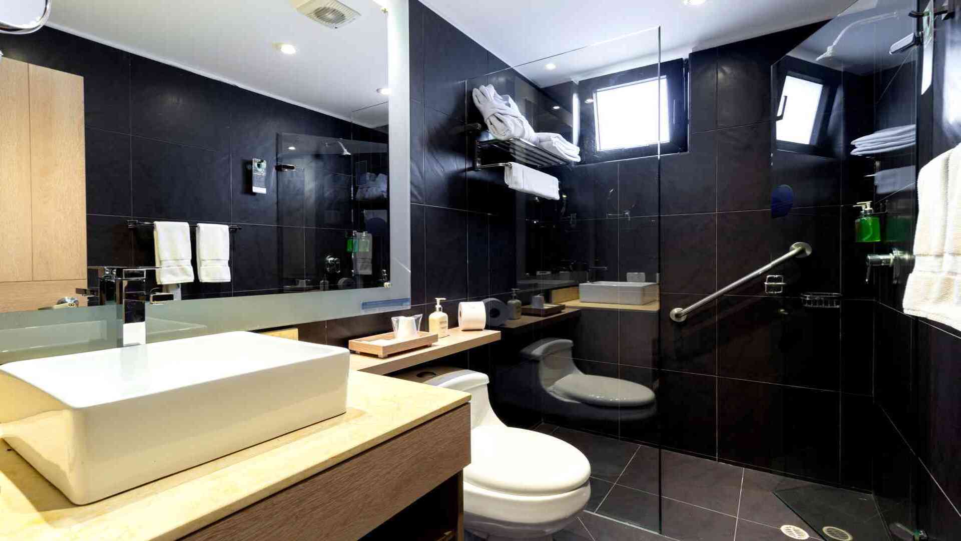 Un baño moderno con azulejos oscuros, lavabo rectangular sobre encimera de madera, inodoro y una ducha. El diseño es elegante y funcional.