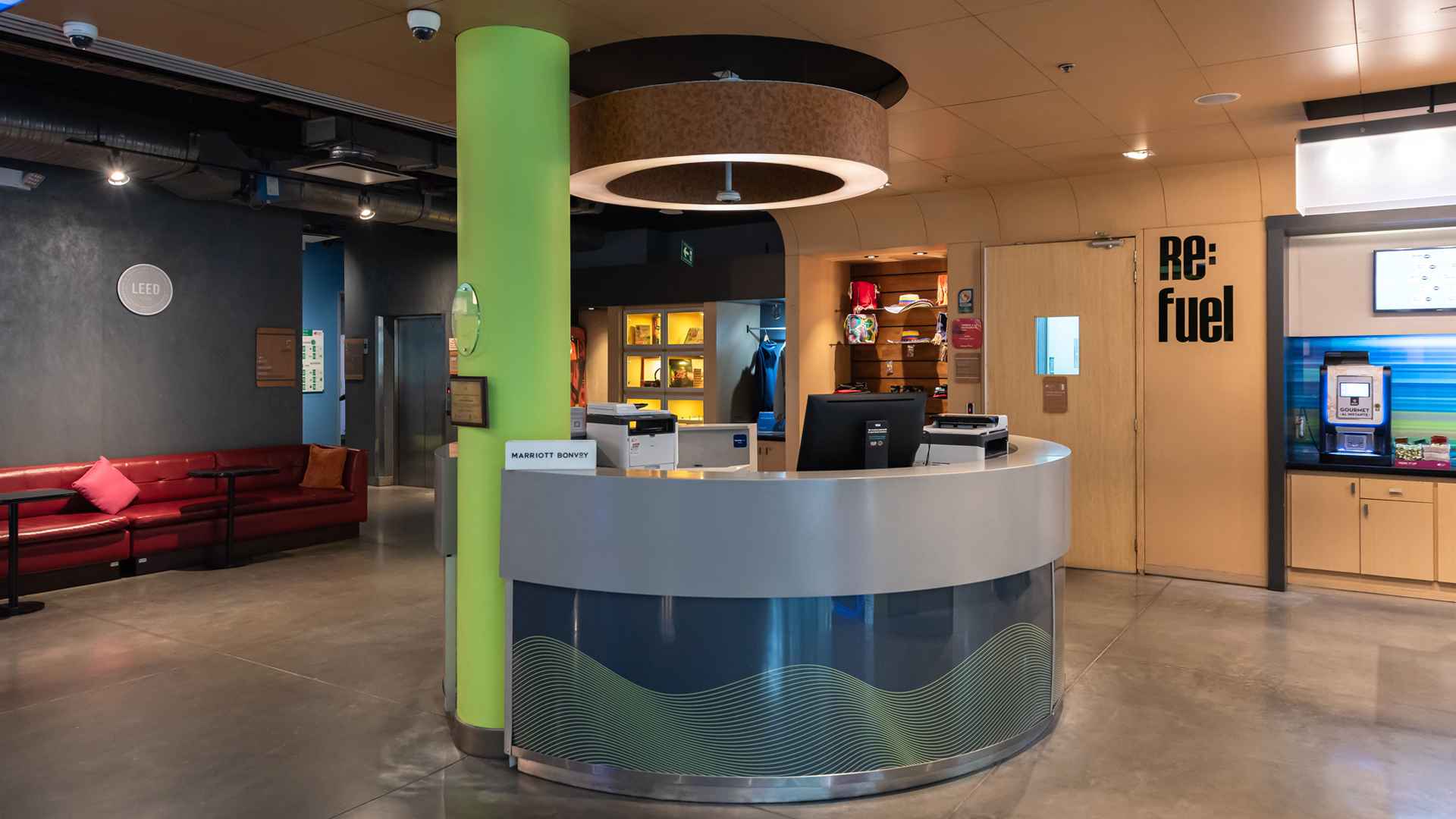 El área de recepción de un hotel Aloft con un mostrador curvo, una columna verde y acceso a la estación "Re:fuel" y un lounge.