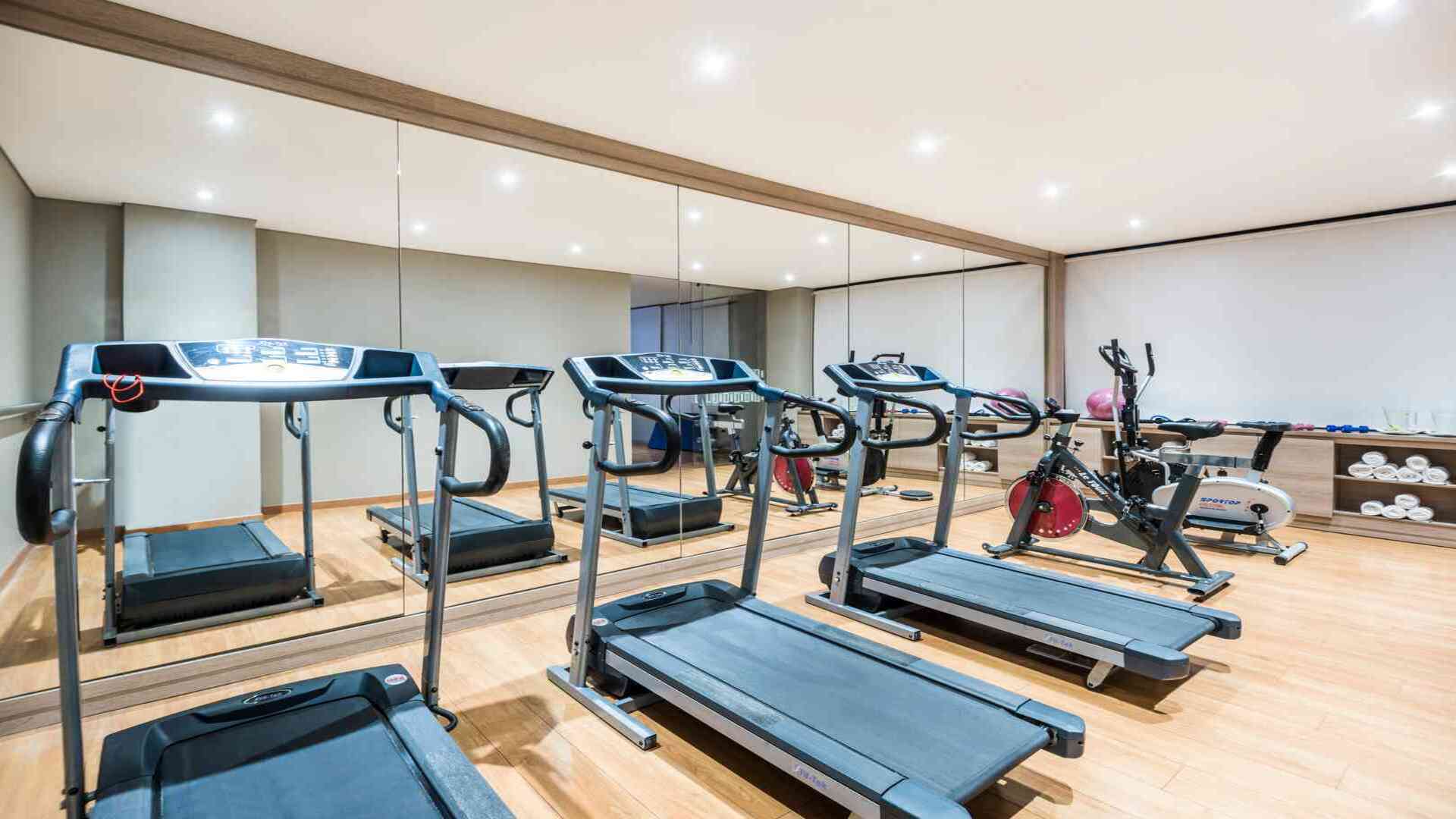 Un gimnasio de hotel con espejos en una pared, equipado con cintas de correr y bicicletas estáticas. El espacio es luminoso y ordenado.
