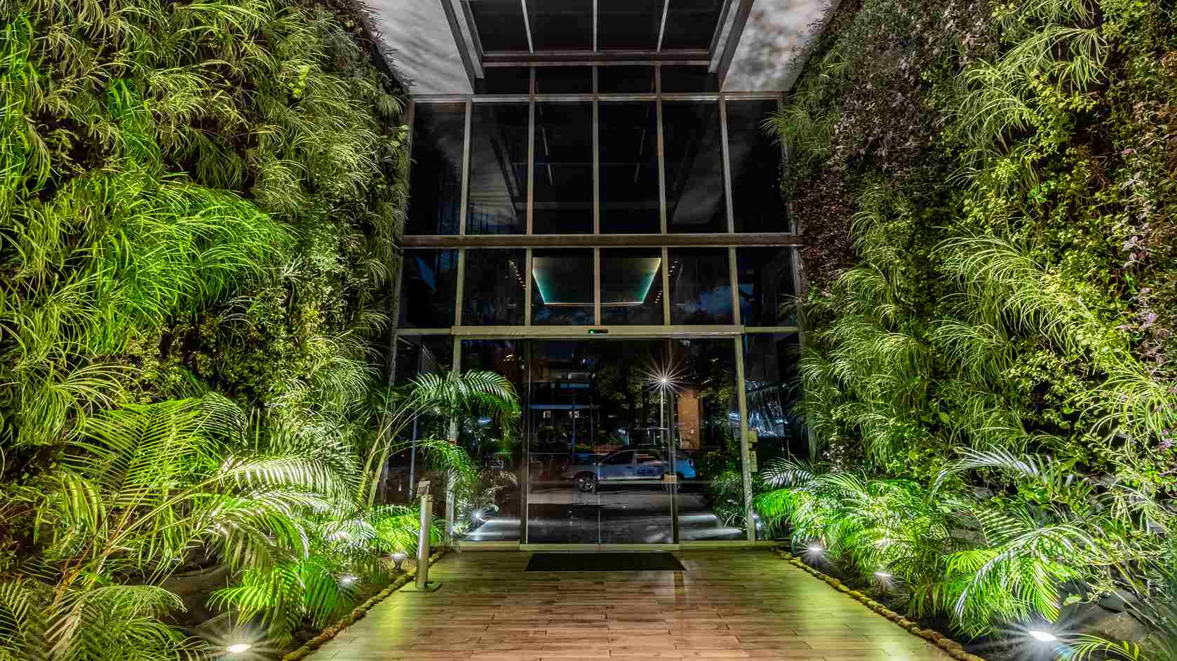 Una entrada espectacular con paredes de vegetación vertical a ambos lados y un pasillo de madera que conduce a puertas de cristal. Iluminación estratégica realza el verdor.