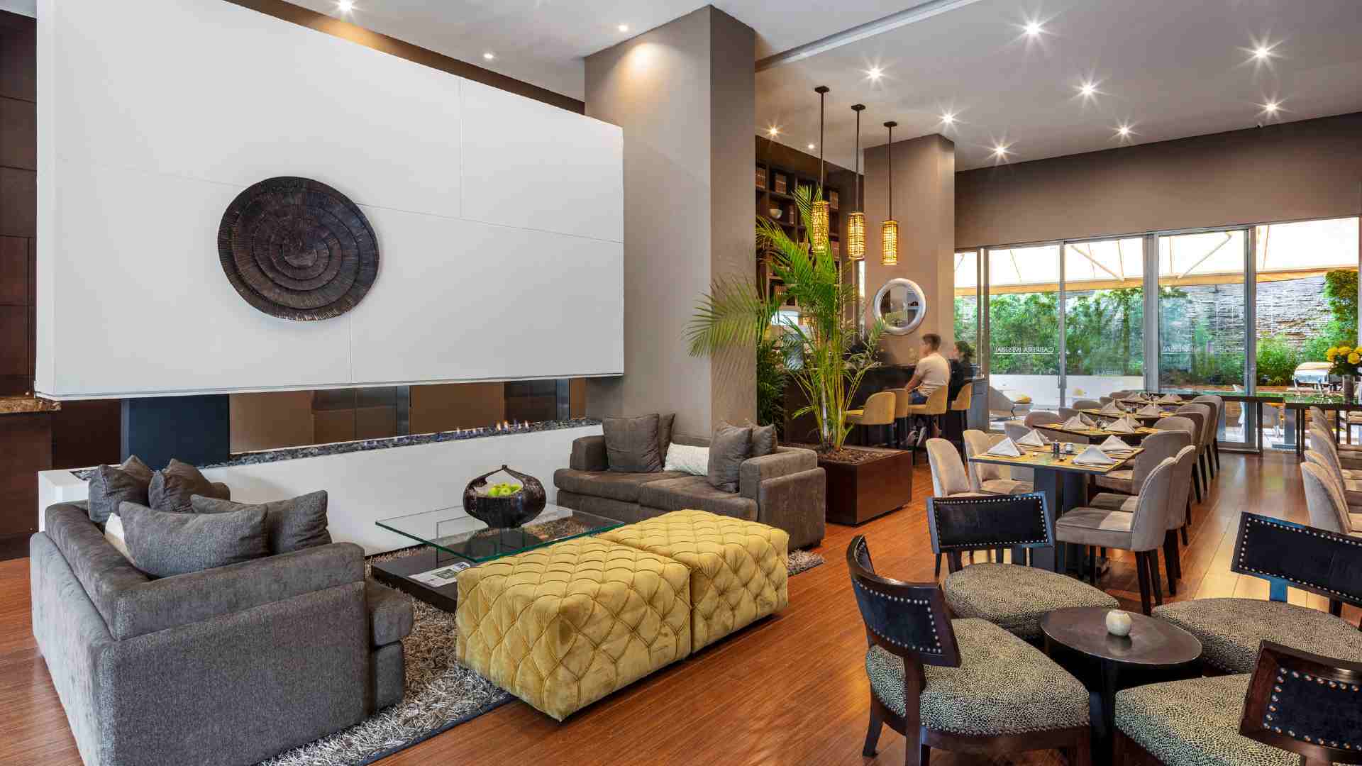 Un moderno lobby lounge que combina un área de sofás frente a una chimenea y un comedor. La decoración es sofisticada, con toques de verde y madera.