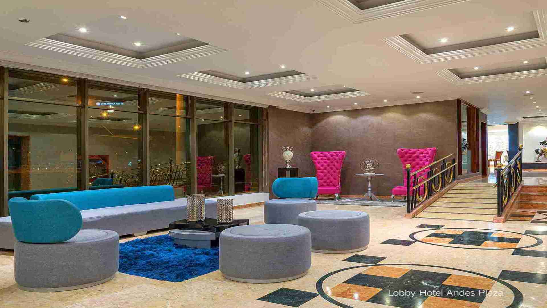 El lobby vibrante del "Hotel Andes Plaza" con sofás azules y pufs grises, contrastando con sillones fucsias. El suelo de mármol tiene diseños geométricos.