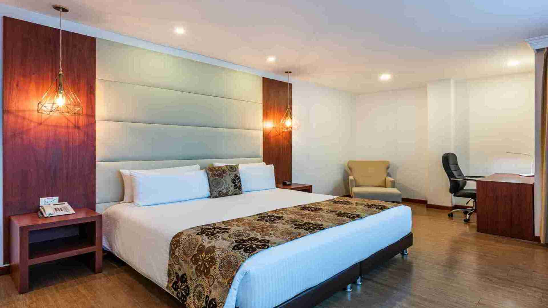  Una habitación de hotel elegante con una cama king-size, cabecera acolchada, detalles en madera y un escritorio con silla. La iluminación colgante añade calidez.