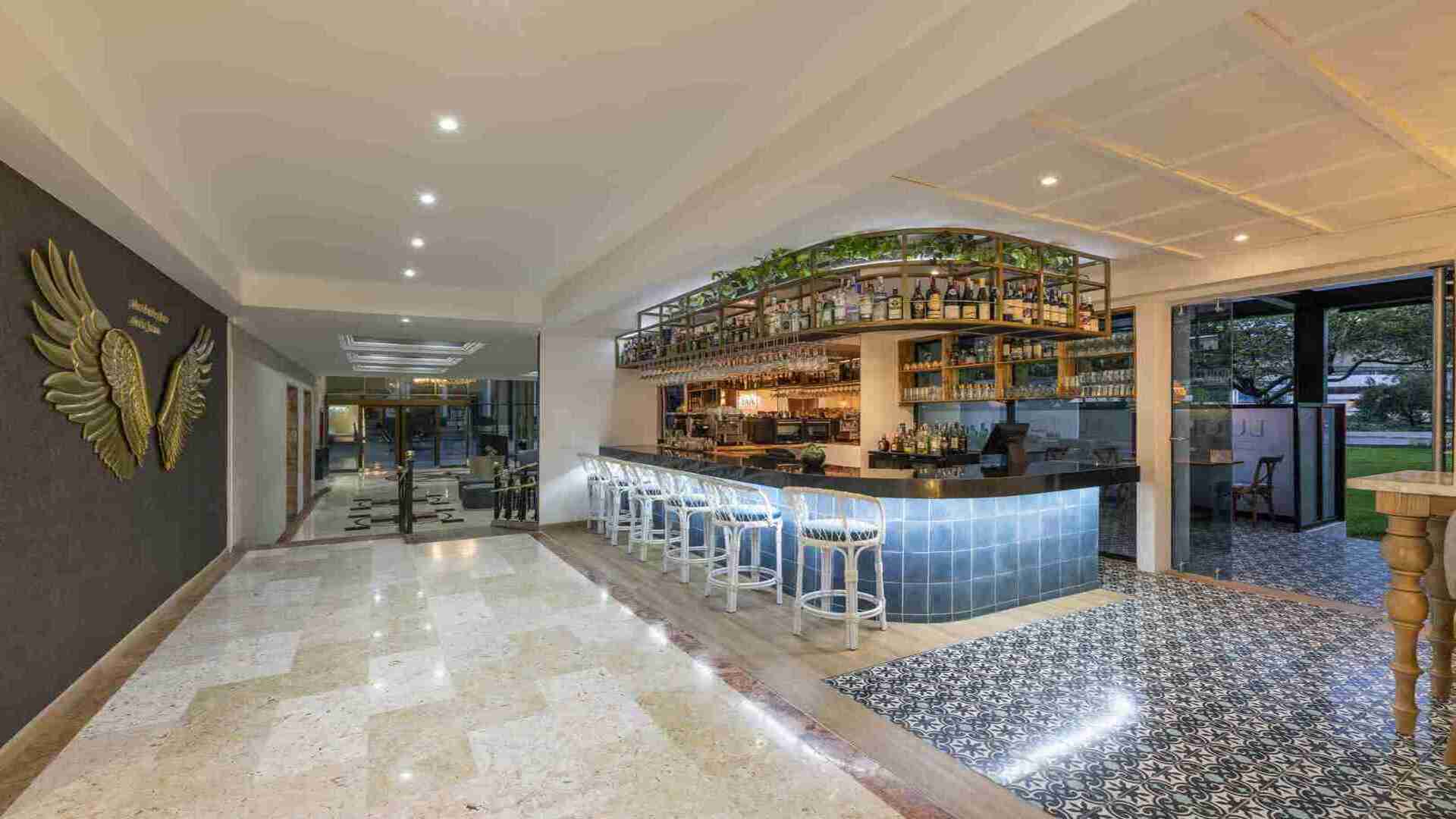 Un bar moderno y sofisticado con una barra iluminada, taburetes blancos y una estantería superior con botellas y plantas. Un espacio ideal para socializar.