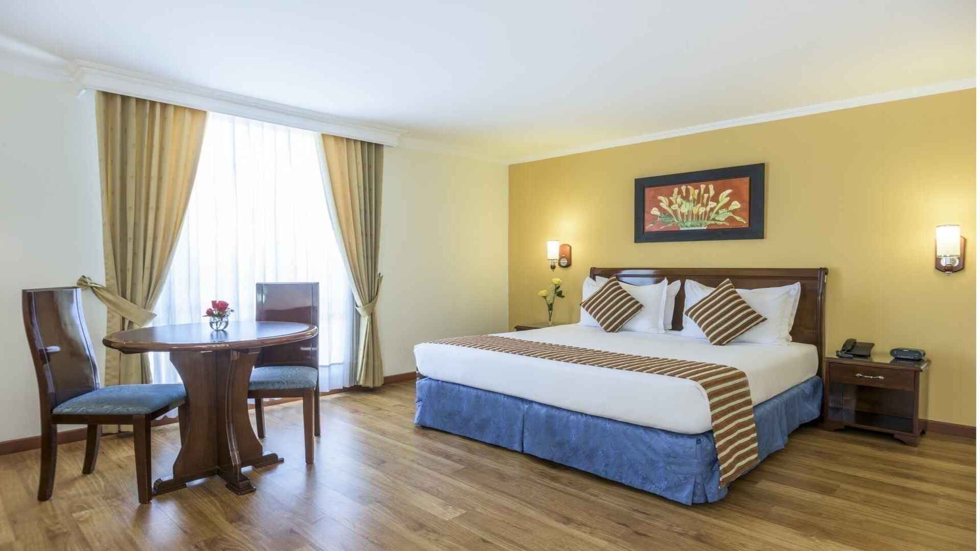  Esta imagen muestra un dormitorio de hotel acogedor con una cama grande, una mesita con dos sillas y una ventana con cortinas. La decoración es clásica con tonos cálidos.