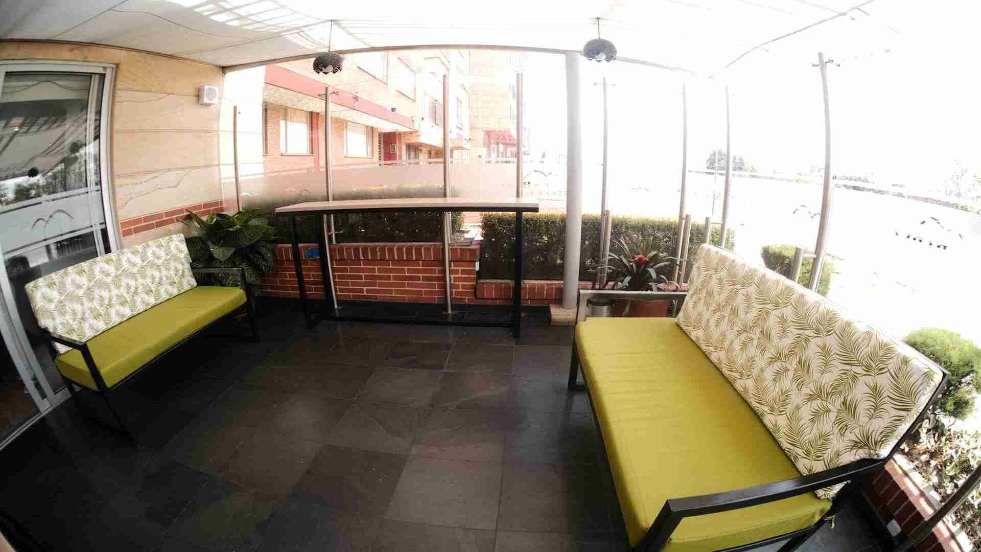 Un área de patio exterior techada y cerrada, con dos bancos con cojines verdes estampados y una mesa alta, ideal para relajarse al aire libre.
