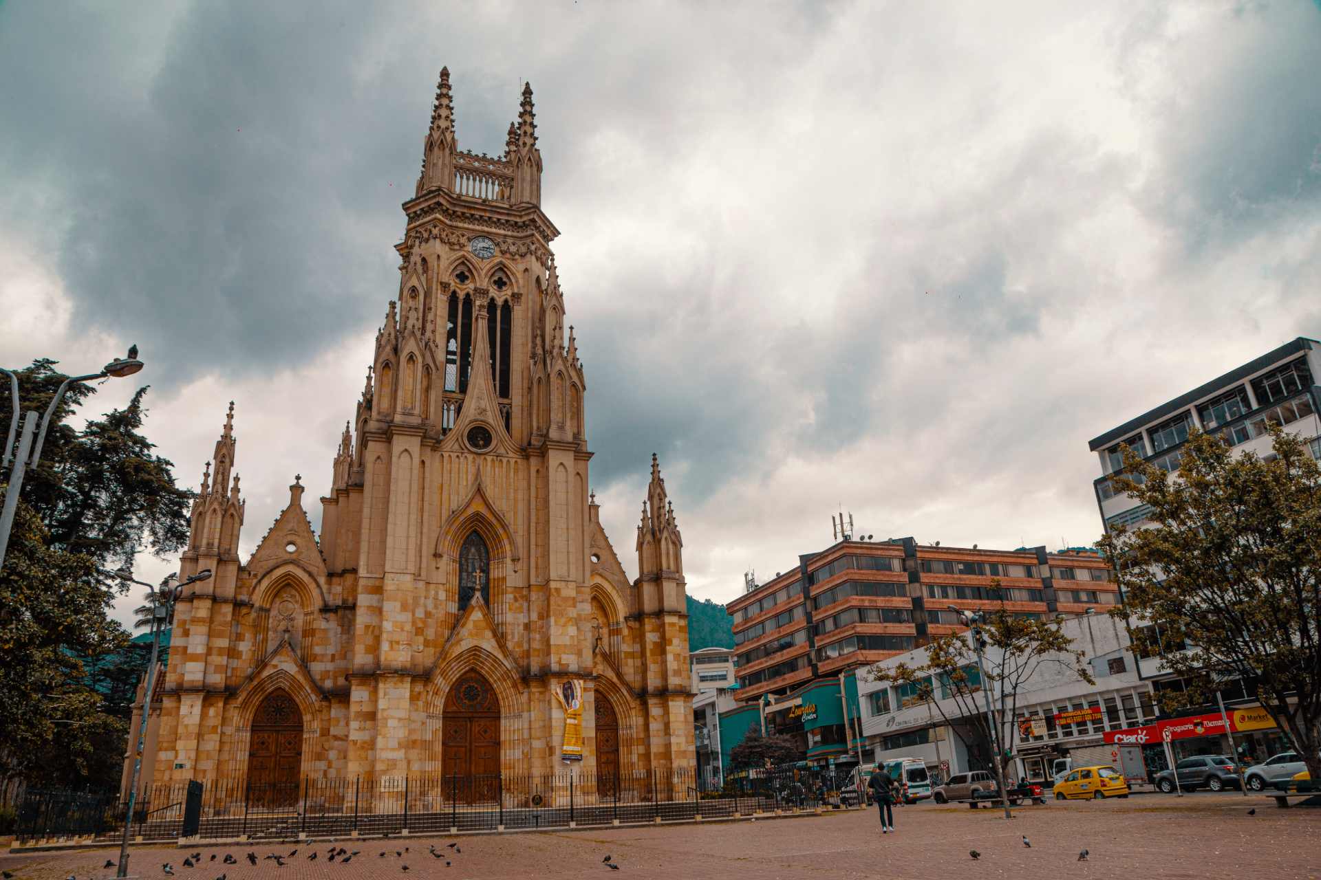  Iglesia de Lourdes de día de arquitectura estilo neogótico, paredes color blanco con detalles en piedra. La iglesia incluye altos arcos y vidrieras. Al frente está la plazoleta vacía con palomas y al rededor centros comerciales.