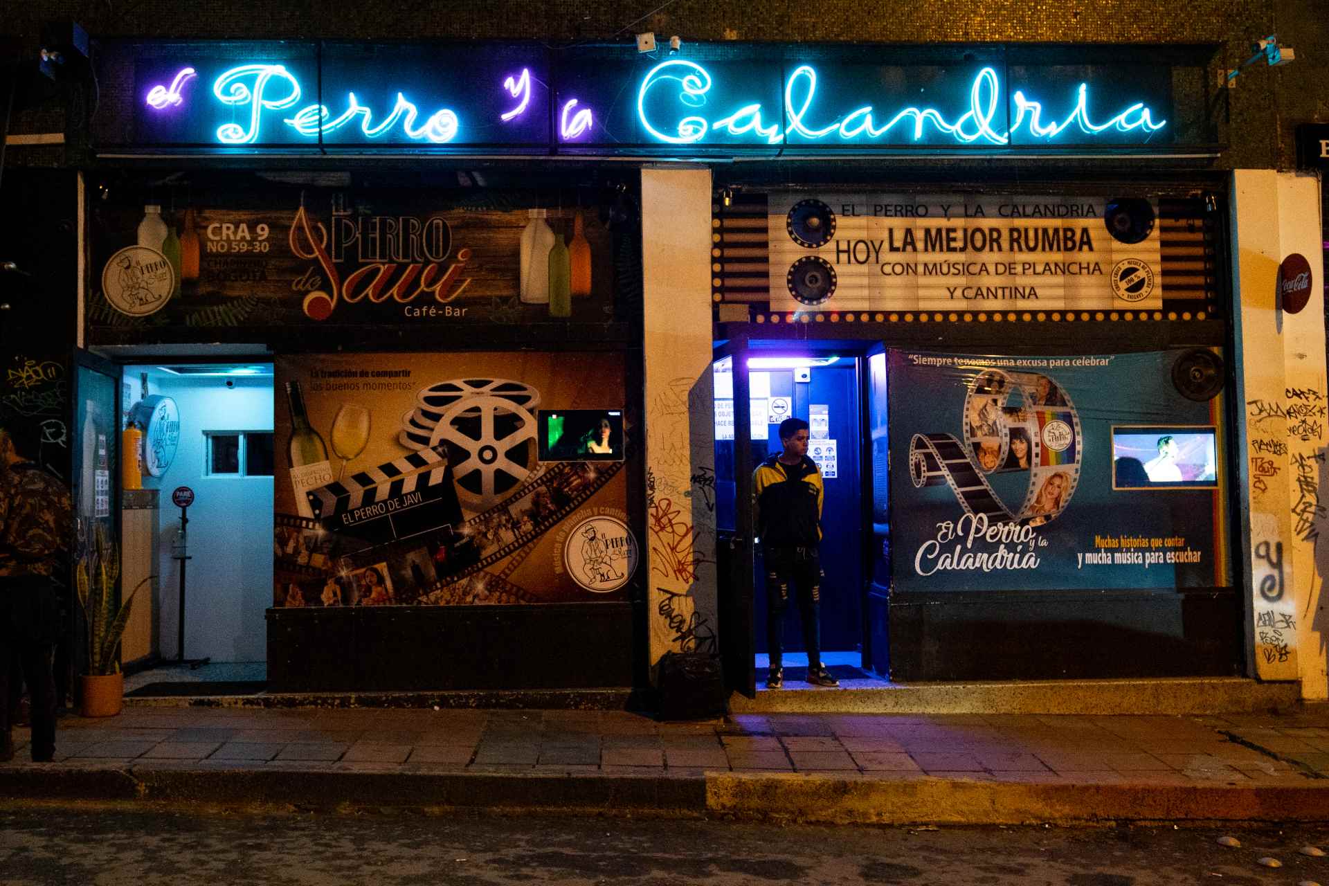 El Perro y la Calandria fachada de noche con aviso en neón color azul, con dos puertas abiertas, un hombre de pie en una de ellas, y la pared cubierta con dos avisos con forma de cartelera de cine.