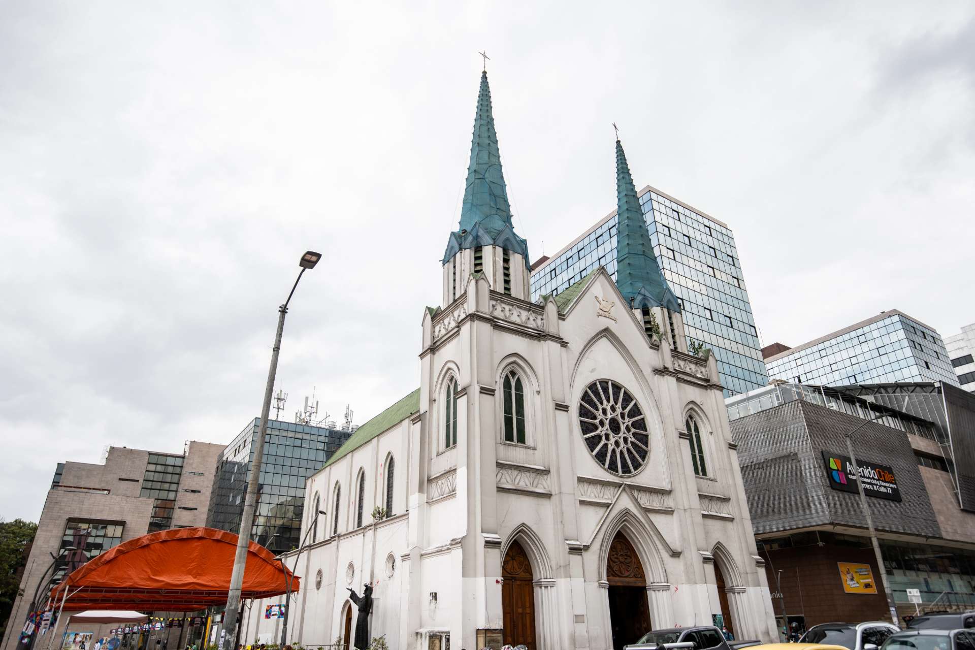 Iglesia de La Porciúncula de día fachada blanca estilo neogótico, con techo de dos agujas de color azul. La rodea edificio moderno que contrasta con su antiguedad. A la izquierda hay una carpa anaranjada de algún evento