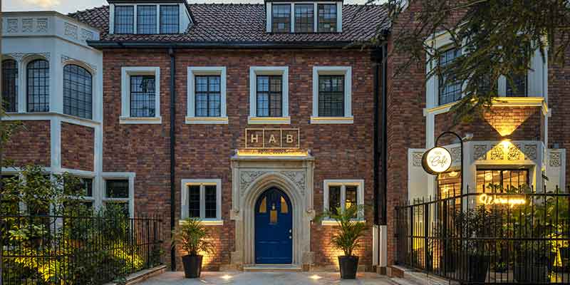 Fachada Hotel HAB al atardecer, casa antigua tipo inglés en ladrillo con 2 antejardines y tres pisos. Puerta principal de madera azul en forma de óvalo con dos materas, una al lado izquirdo y otra al derecho. De una ventana sale luz artificial.
