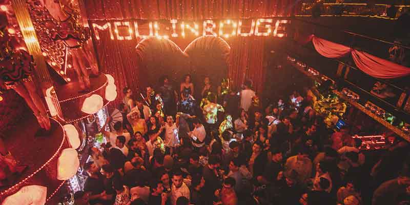 Chiquita bar interior con multitud de personas bailando. Se ve un aviso de luz blanca que dice Moulin Rouge.
