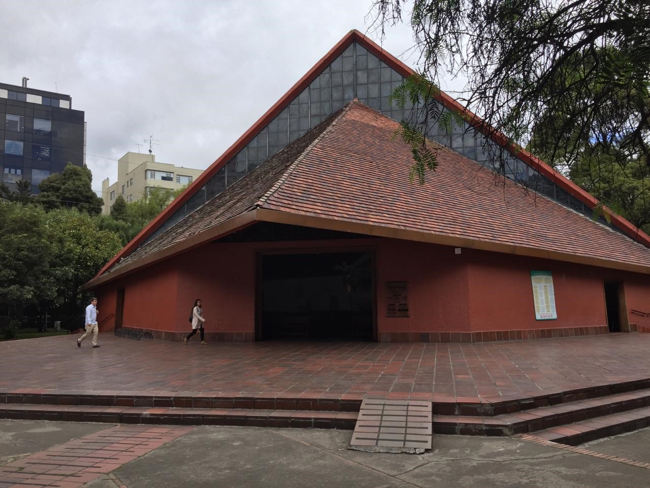  Iglesia Inmaculada Concepción, fachada de día con cielo gris. Estructura triangular en cemento rojo, ladrillo y grandes ventanales en forma de cuadrícula. Por el frente en baldosa de ladrillo caminan un hombre y una mujer. Mientras al frente están las ramas de un árbol al fondo destaca un edificio moderno y árboles frondosos.