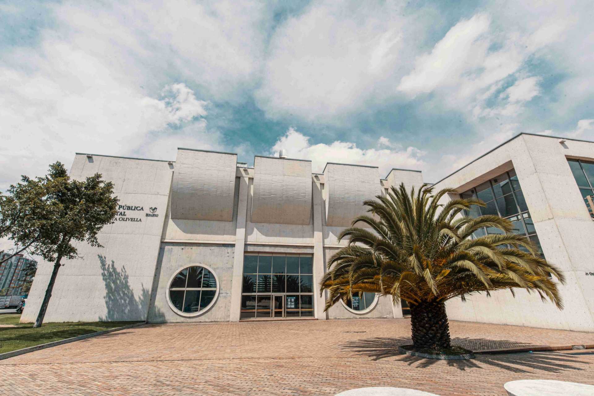 Biblioteca El Tintal fachada de día paredes en concreto blanco con grandes ventanales circulares y cuadrados y gran puerta cuadrada en vidrio. Al frente a la derecha hay una palma de mediano tamaño y a la izquierda un árbol frondoso.  El cielo es azul con nubes blancas.