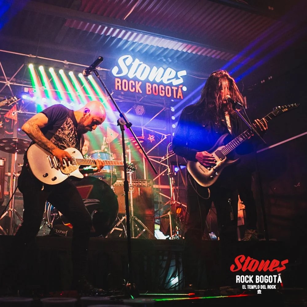 Escenario de banda de rock con aviso de color blanco y rojo con eslogan de Stones Rock Bogotá. Sobresalen un cantante de cabello largo y un guitarrista calvo. Al fondo se ve un baterista.